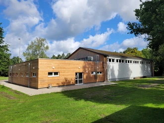 Sporthalle_in_Huetzel_01.jpg Sporthalle in Hützel mit Holzfassade und angrenzendem weißem Hallenbereich unter bewölktem Himmel