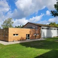 Sporthalle_in_Huetzel_01.jpg Sporthalle in Hützel mit Holzfassade und angrenzendem weißem Hallenbereich unter bewölktem Himmel