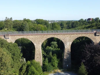 Saubrücke_Eulenbachbrücke_02_Stadtmarketing Velbert.JPG Eine historische Steinbrücke mit fünf Bögen überspannt ein bewaldetes Tal in einer grünen Umgebung.