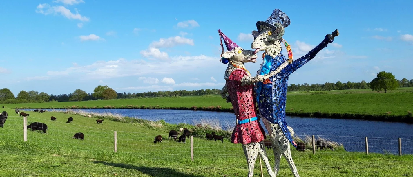 Tanz der Schafe-Vreschen Bokel-Apen-Foto Sandra Hinrichs.jpg Zwei bunte Schafskulpturen tanzen auf einer Weide am Fluss, dahinter grasen echte Schafe.
