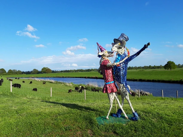 Tanz der Schafe-Vreschen Bokel-Apen-Foto Sandra Hinrichs.jpg Zwei bunte Schafskulpturen tanzen auf einer Weide am Fluss, dahinter grasen echte Schafe.