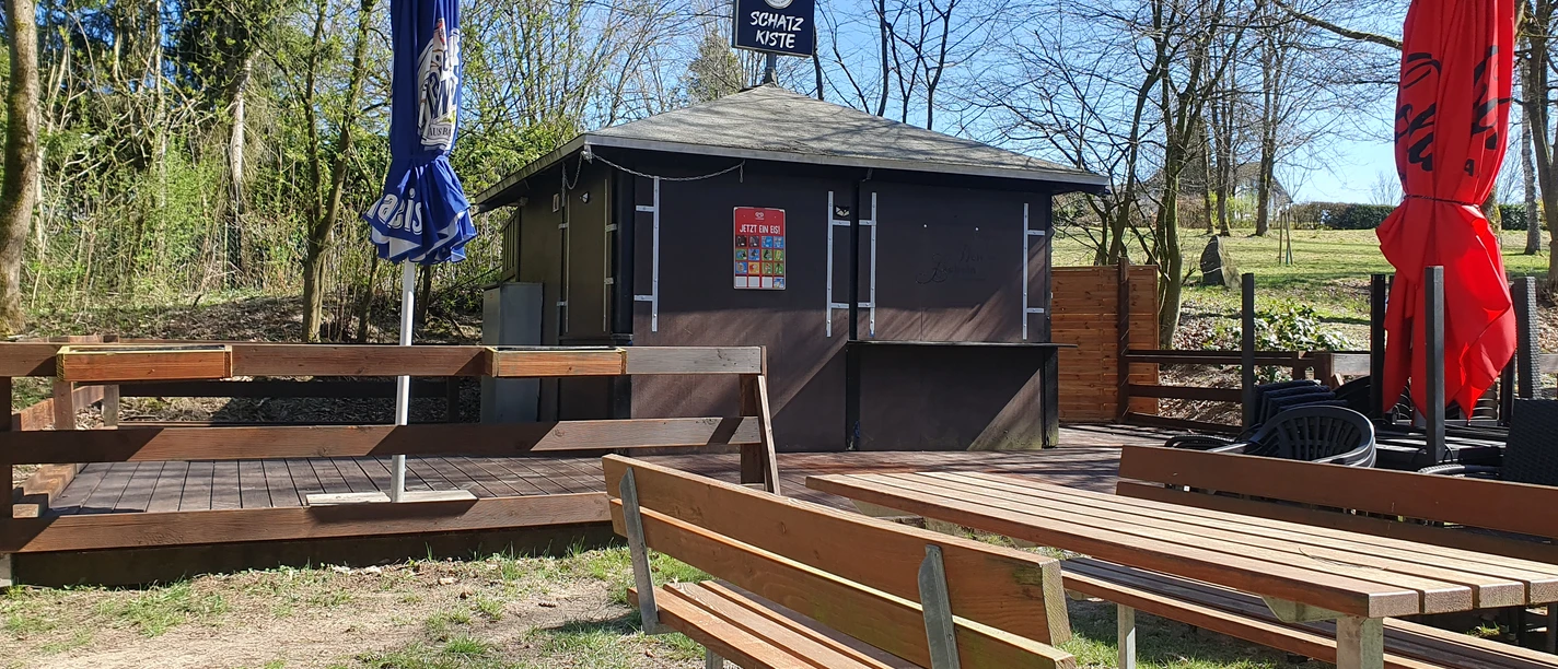 Holzkiosk mit Schiebeläden und Sonnenschirmen, umgeben von Bäumen, wartet auf Gäste.