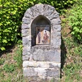Eine steinerne Wegkapelle am Wegesrand, umgeben von grünem Laub und Gras. In einer Nische ist eine religiöse Darstellung angebracht, die eine Szene mit zwei Figuren zeigt – eine stehende Gestalt in weißem Gewand und eine sitzende Figur in Rot, beide mit Heiligenschein. Die Szene ist Teil des Kreuzwegs am Jakobsberg.