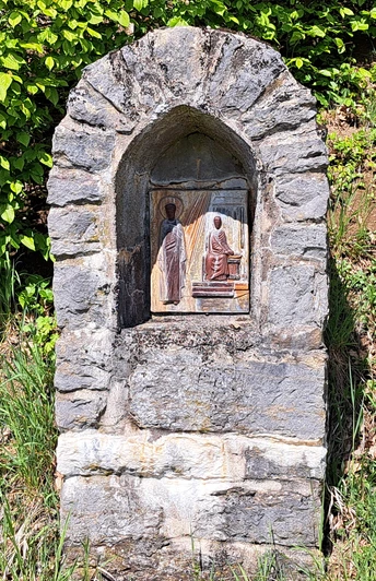 Kreuzwegstation Jakobsberg Eine steinerne Wegkapelle am Wegesrand, umgeben von grünem Laub und Gras. In einer Nische ist eine religiöse Darstellung angebracht, die eine Szene mit zwei Figuren zeigt – eine stehende Gestalt in weißem Gewand und eine sitzende Figur in Rot, beide mit Heiligenschein. Die Szene ist Teil des Kreuzwegs am Jakobsberg.