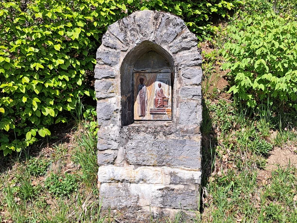 Kreuzwegstation Jakobsberg Eine steinerne Wegkapelle am Wegesrand, umgeben von grünem Laub und Gras. In einer Nische ist eine religiöse Darstellung angebracht, die eine Szene mit zwei Figuren zeigt – eine stehende Gestalt in weißem Gewand und eine sitzende Figur in Rot, beide mit Heiligenschein. Die Szene ist Teil des Kreuzwegs am Jakobsberg.