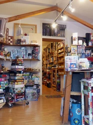 Spielbrett (Innenraum) Innenraum eines gut sortierten Brettspielladens mit einer Fülle an bunten Spieleschachteln, die auf Regalen und Böden gestapelt sind. Eine helle Deckenbeleuchtung sorgt für eine einladende Atmosphäre.