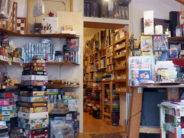 Spielbrett (Innenraum) Innenraum eines gut sortierten Brettspielladens mit einer Fülle an bunten Spieleschachteln, die auf Regalen und Böden gestapelt sind. Eine helle Deckenbeleuchtung sorgt für eine einladende Atmosphäre.