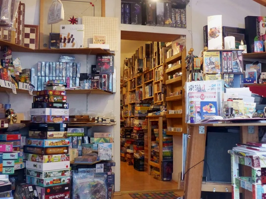 Spielbrett (interior) Innenraum eines gut sortierten Brettspielladens mit einer Fülle an bunten Spieleschachteln, die auf Regalen und Böden gestapelt sind. Eine helle Deckenbeleuchtung sorgt für eine einladende Atmosphäre.Interior of a well-stocked board game store with an abundance of colorful game boxes stacked on shelves and floors. Bright ceiling lighting creates an inviting atmosphere.