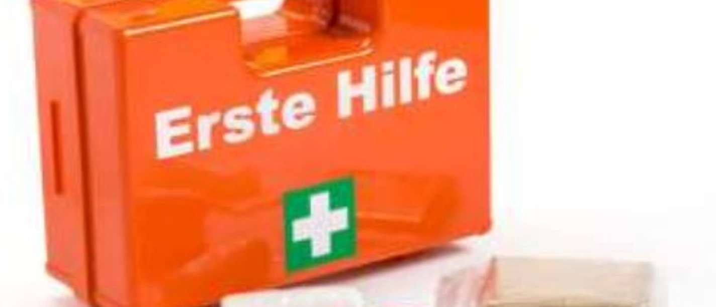 Ein orangefarbener Erste-Hilfe-Kasten mit weißem Text sowie Verbandmaterialien und Schere im Vordergrund.