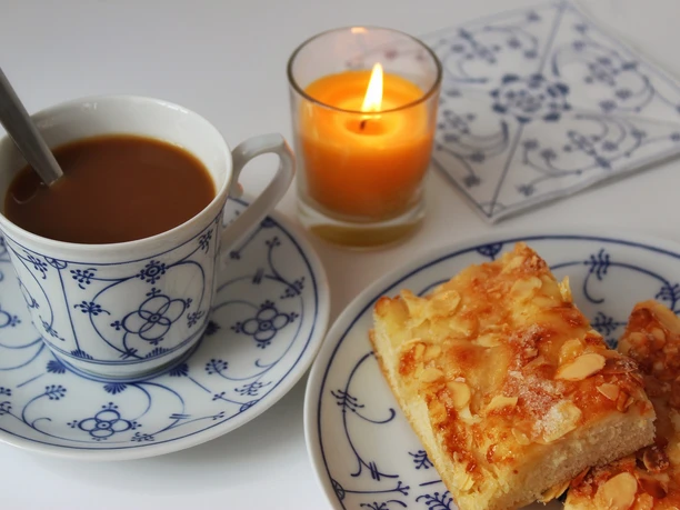 Butterkuchen Ein Stück Butterkuchen mit gerösteten Mandeln liegt auf einem Teller neben einer Tasse Kaffee und einer brennenden Kerze.