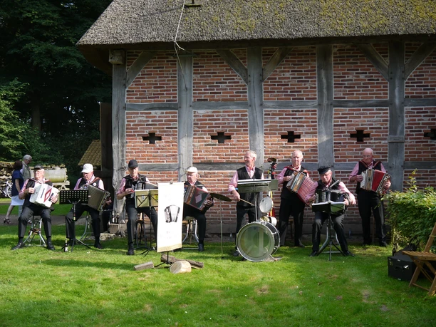 musikgruppe-im-freilichtmuseum.JPG Eine Gruppe älterer Männer spielt Akkordeon in traditioneller Kleidung vor einem alten Gebäude auf einer Wiese.