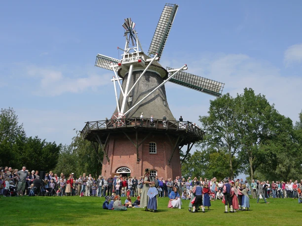 tag-der-heimat-bei-der-muehle.jpg Menschen in traditionellen Gewändern versammeln sich vor einer historischen Mühle bei einer Feier.