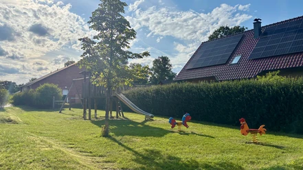 Spielplatz Bloumstücken.jpg Spielplatz Bloumstücken mit Klettergerüst und Rutsche, zwei Federtiere auf Grasfläche, Haus mit Solarpaneelen im Hintergrund