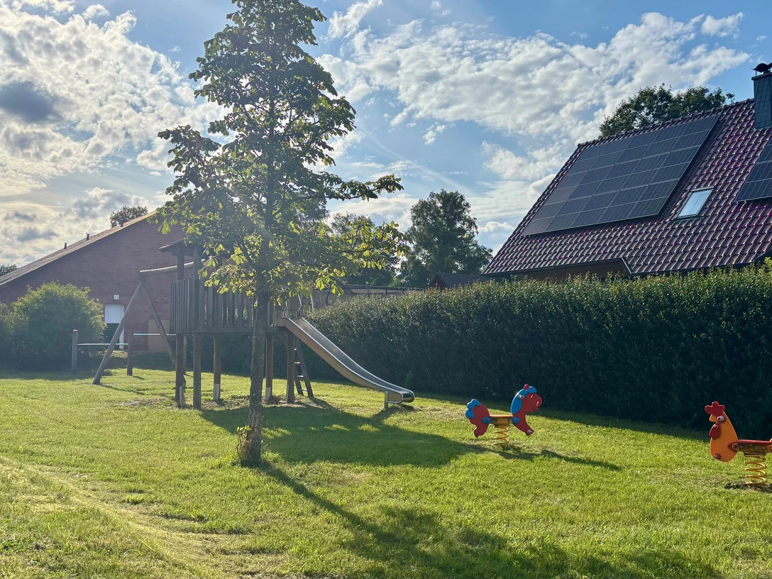Spielplatz Bloumstücken.jpg Spielplatz Bloumstücken mit Klettergerüst und Rutsche, zwei Federtiere auf Grasfläche, Haus mit Solarpaneelen im Hintergrund