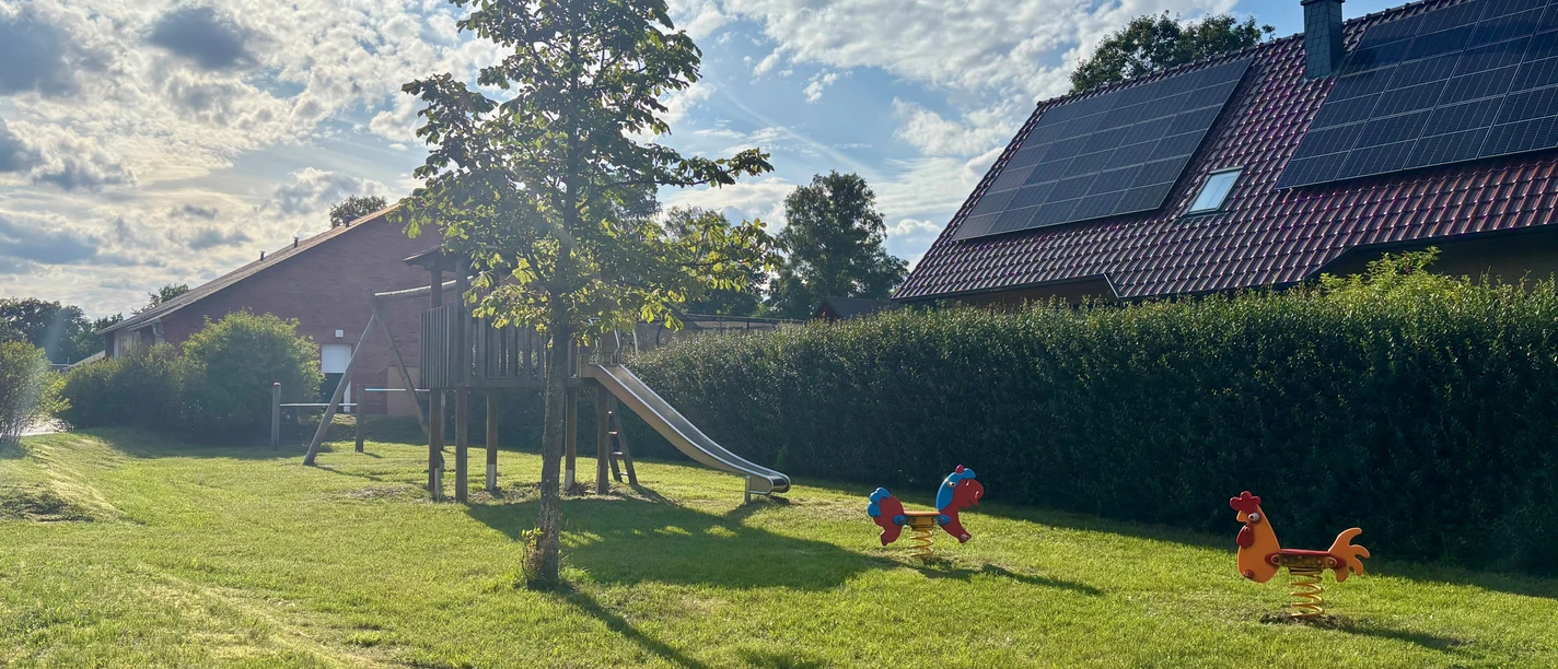 Spielplatz Bloumstücken.jpg Spielplatz Bloumstücken mit Klettergerüst und Rutsche, zwei Federtiere auf Grasfläche, Haus mit Solarpaneelen im Hintergrund