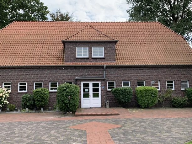 Dörpshus Bokeler School Das rote Backsteingebäude mit Satteldach hat eine zentral gelegene, weiße Eingangstür und kleine Fenster. Eine Reihe Büsche sind vor dem Haus gepflanzt.Davor befindet sich eine gepflasterte Fläche.