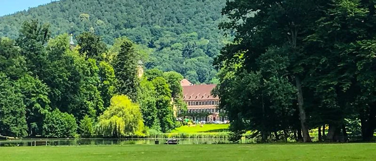 Fürstliche Abtei Idyllische Landschaft mit grüner Wiese, See und einer fürstlichen Abtei vor bewaldetem Hügel.