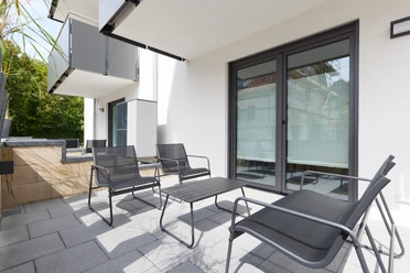 Terrasse mit modernen Metallstühlen und -tisch, umringt von weißen Gebäudewänden, darunter Glasfenster.