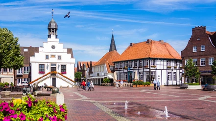Historisches Rathaus mit Petunien - Foto Helmut Kramer 170703111111.jpg Weißes Rathaus mit Treppen und Blumentöpfen auf dem Marktplatz von Lingen bei blauem Himmel.