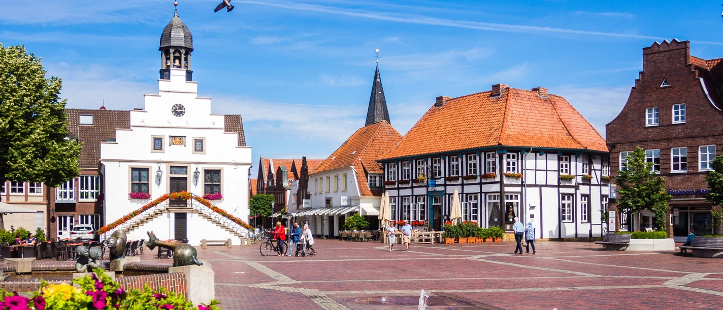 Historisches Rathaus mit Petunien - Foto Helmut Kramer 170703111111.jpg Weißes Rathaus mit Treppen und Blumentöpfen auf dem Marktplatz von Lingen bei blauem Himmel.