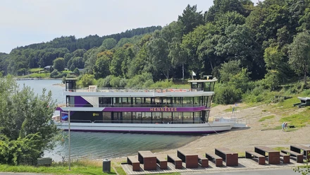 EMS Hennesee Ein weißes Ausflugsschiff mit zwei Decks liegt an einem bewaldeten Ufer des Hennesees an.