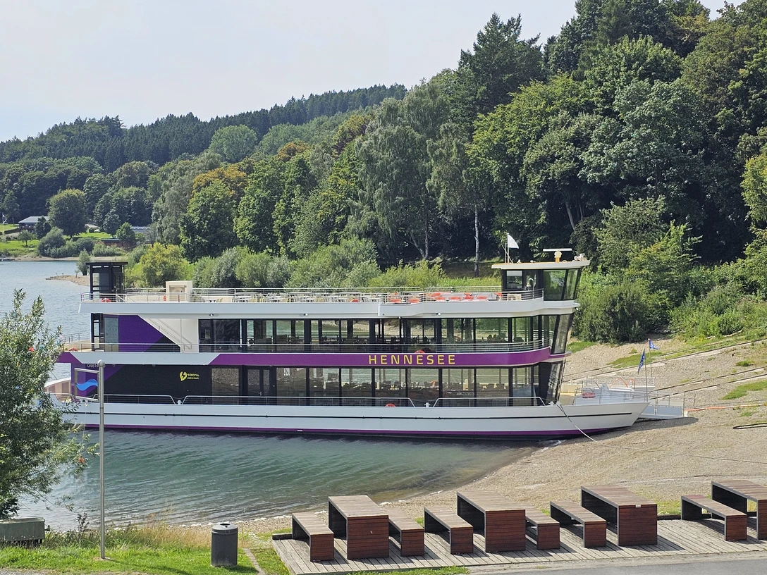 EMS Hennesee Ein weißes Ausflugsschiff mit zwei Decks liegt an einem bewaldeten Ufer des Hennesees an.