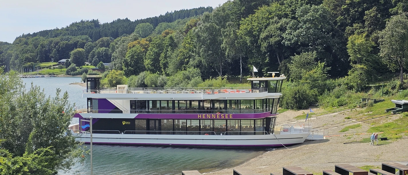EMS Hennesee Ein weißes Ausflugsschiff mit zwei Decks liegt an einem bewaldeten Ufer des Hennesees an.