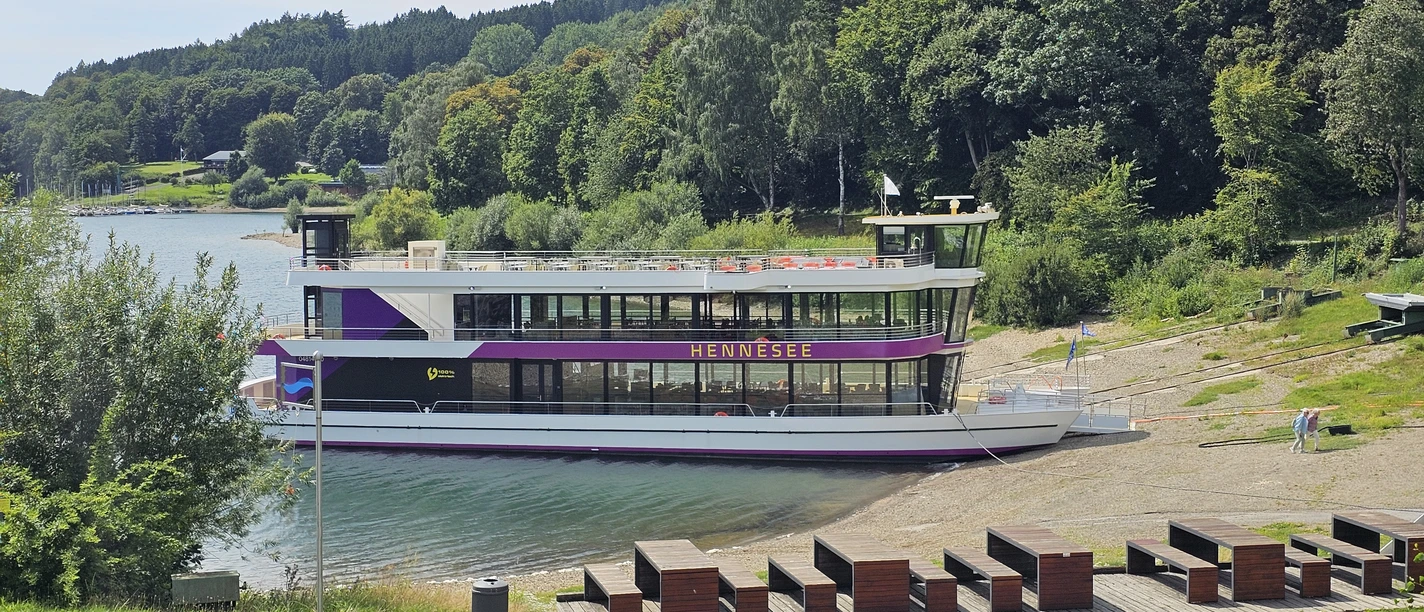 Ein weißes Ausflugsschiff mit zwei Decks liegt an einem bewaldeten Ufer des Hennesees an.