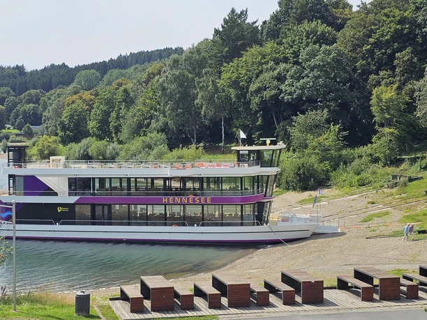 EMS Hennesee Ein weißes Ausflugsschiff mit zwei Decks liegt an einem bewaldeten Ufer des Hennesees an.