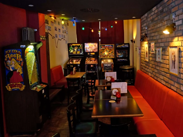 Spiel- und Flipperautomaten in der Red Fox Bar Das Bild zeigt eine Bar mit dunklem Holzmobiliar, roten Sitzbänken und mehreren Flipperautomaten an der Wand. Bunte Lichterketten hängen von der Decke, während an den Wänden verschiedene Bilder und Retro-Dekorationen zu sehen sind.