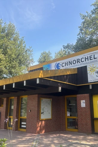 IMG_2889.jpg Backsteingebäude mit gelben Türrahmen und flachem Dach, umgeben von Bäumen und Sonnenschein.