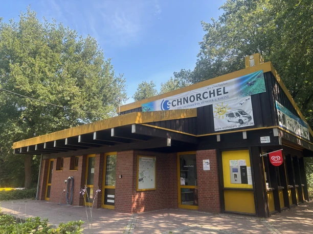 IMG_2889.jpg Backsteingebäude mit gelben Türrahmen und flachem Dach, umgeben von Bäumen und Sonnenschein.