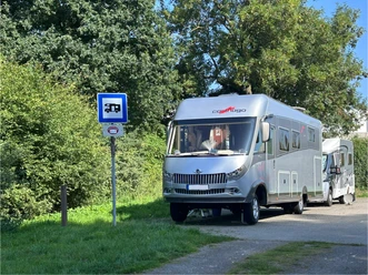 Zwei geparkte Wohnmobile auf befestigtem Stellplatz am Sportzentrum, umgeben von Bäumen und Grünflächen.