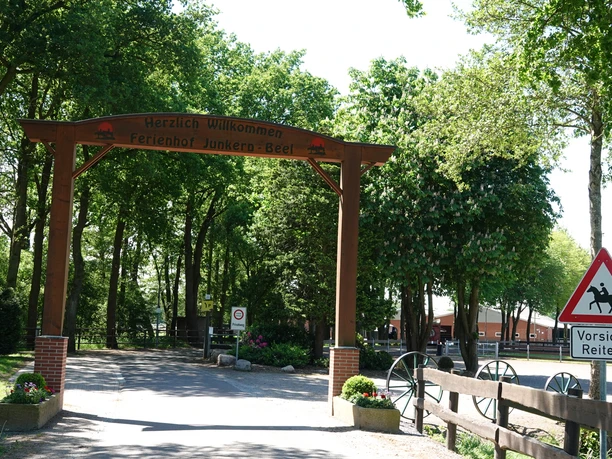De entree van vakantieboerderij Junkern-Beel met houten poort, waarschuwingsbord voor ruiters en bomen eromheen.
