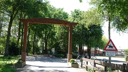 Ferienhof Junkern-Beel, Lathen-Hilter ©Emsland Tourismus GmbH (3).JPG Eingangsbereich des Ferienhofs Junkern-Beel mit Holzportal, Reiterwarnschild und umgebenden Bäumen.
