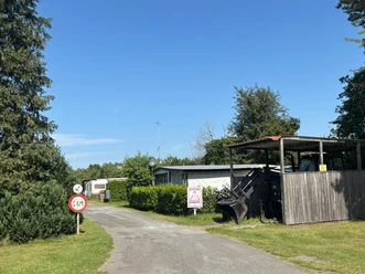 Schmaler Zufahrtsweg mit Schild, Wohnwagen und Traktor neben einem Holzunterstand unter blauem Himmel.