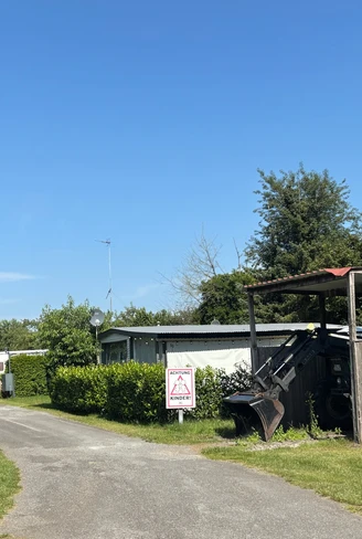 Schmaler Zufahrtsweg mit Schild, Wohnwagen und Traktor neben einem Holzunterstand unter blauem Himmel.