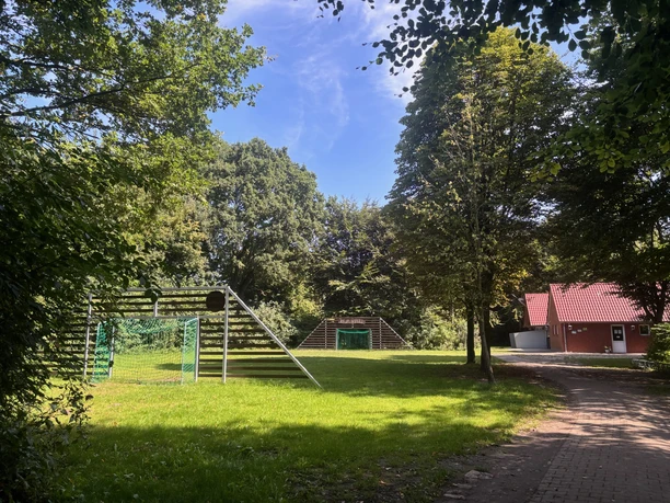 Jugendzeltplatz Vrees | 1 Grüne Wiese mit Fußballtoren und Holzkletterwand, umgeben von Bäumen, neben rotem Gebäude mit Dach.
