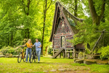 Batakhaus, Werpeloh – Radfahren Hümmling ©Emsland Tourismus GmbH (3).jpg Zwei Radfahrende vor dem traditionellen Batakhaus in Werpeloh, umgeben von grünem Laubwald.