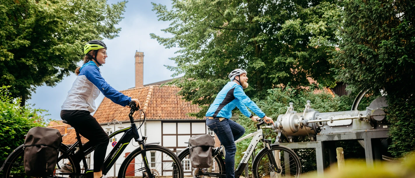 Am Tuchmacher Museum in Bramsche Zwei Radfahrer mit Helmen fahren auf einem gepflasterten Weg an einem Fachwerkhaus vorbei.