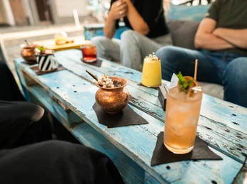 Toddy Tapper Das Bild zeigt eine Gruppe von Menschen, die draußen an einem Tisch aus blauen Holzbrettern sitzen. Vor ihnen stehen dekorative Getränke in verschiedenen Gläsern. Es herrscht eine entspannte Atmosphäre.The picture shows a group of people sitting outside at a table made of blue wooden boards. In front of them are decorative drinks in various glasses. The atmosphere is relaxed.