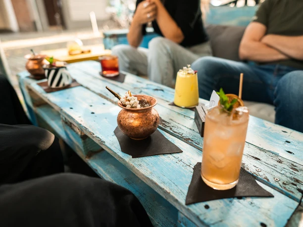 Toddy Tapper The picture shows a group of people sitting outside at a table made of blue wooden boards. In front of them are decorative drinks in various glasses. The atmosphere is relaxed.