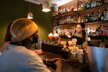 Toddy Tapper Eine Barkeeperin mixt Cocktails in einer atmosphärischen Bar vor aufmerksamem Publikum.