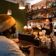 Toddy Tapper Eine Barkeeperin mixt Cocktails in einer atmosphärischen Bar vor aufmerksamem Publikum.