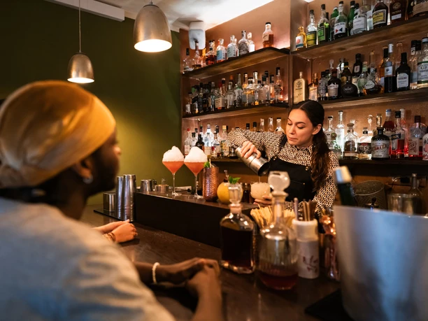 Toddy Tapper A bartender mixes cocktails in an atmospheric bar in front of an attentive audience.