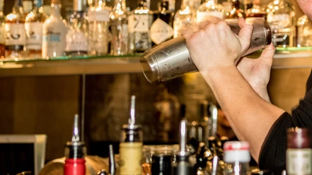 Bar Ein Barkeeper schüttelt einen Edelstahl-Shaker, im Hintergrund sind Flaschen zu sehen.