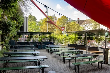 Zum Scheuen Reh Grüner Biergarten mit Holzbänken und Tischen, eingerahmt von Pflanzen, unter blauem Himmel.