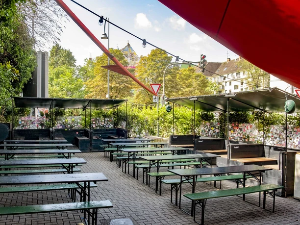 Zum Scheuen Reh Grüner Biergarten mit Holzbänken und Tischen, eingerahmt von Pflanzen, unter blauem Himmel.