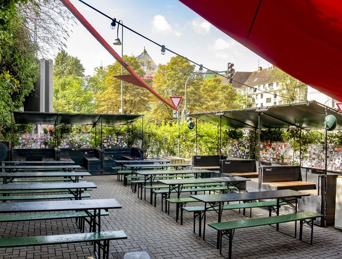 Zum Scheuen Reh Grüner Biergarten mit Holzbänken und Tischen, eingerahmt von Pflanzen, unter blauem Himmel.Green beer garden with wooden benches and tables, framed by plants, under a blue sky.