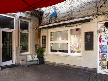 Zum Scheuen Reh Das Bild zeigt einen Außenbereich mit einer weiß gestrichenen Wand, auf der eine stilisierte Hirschfigur angebracht ist. Der Eingang in einem urbanen Straßenkontext mit Plakaten auf der Tür bietet eine einladende Atmosphäre.The picture shows an outdoor area with a white-painted wall on which a stylized deer figure is mounted. The entrance in an urban street context with posters on the door offers an inviting atmosphere.